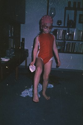 Child in vintage red swimwear and goggles stands indoors, likely a poolside changing area. Notebooks and glassware on shelves...