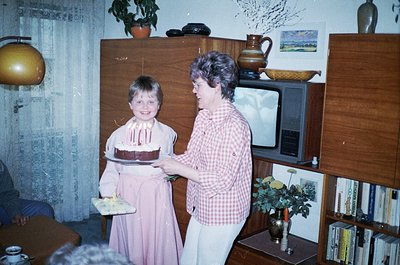 Vintage indoor birthday scene featuring a young girl in a pastel dress holding a round chocolate cake with candles, presented...