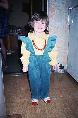 Child in vintage 1970s-80s overalls with ruffled yellow top and red beaded necklace, standing on wooden floor. Reflective red...