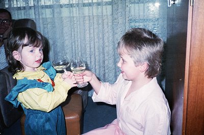 Two children in 1970s-style clothing toast indoors with wine glasses. Girl in a yellow-and-blue patterned dress with a red co...