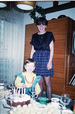 Vintage indoor birthday scene: woman in 1970s-style navy blouse & plaid skirt poses with young girl in yellow top & red beade...