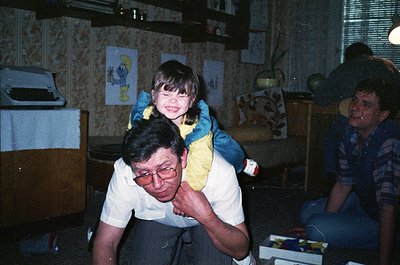 Vintage indoor family moment: man in glasses carries a smiling child in a yellow sweater, another adult watches. Patterned wa...