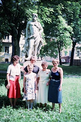 Five women pose outdoors beside a neoclassical statue in a park setting, likely Eastern Europe, 1960s–1970s. The woman in the...