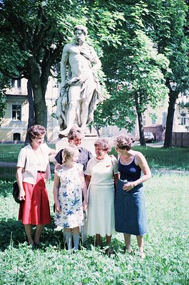 Classic 1960s group portrait in a European park, featuring six adults posing beside a neoclassical statue. Women wear floral ...