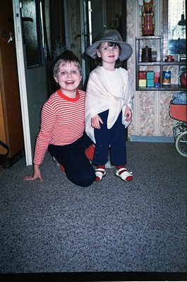 Two children pose indoors, likely mid-1980s–90s. The boy kneels in a striped red-and-white sweater and dark pants, while the ...