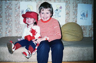 Two children pose indoors on a patterned sofa, likely 1980s–1990s. The boy wears a red-and-white striped sweater, blue jeans,...