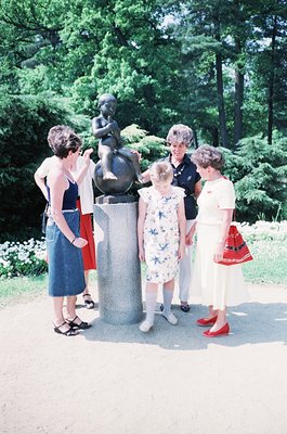 Four women pose beside a bronze statue of a seated child in a lush park setting, likely mid-20th century. The woman in the ce...
