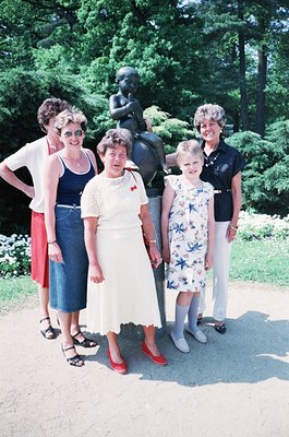 Four women pose outdoors beside a bronze statue of a seated child in a park setting, likely mid-20th century. The central wom...