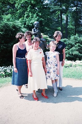 Five women pose outdoors in a landscaped park setting, likely mid-20th century (1960s–1970s). Central figure wears a white dr...