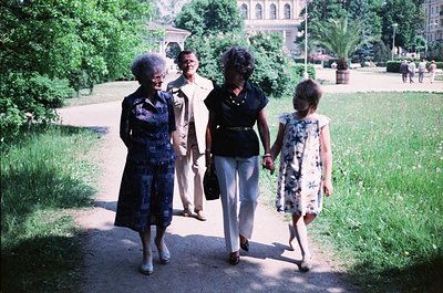 Four women stroll through a landscaped park with manicured lawns and trees, likely mid-20th century. The woman on the right w...