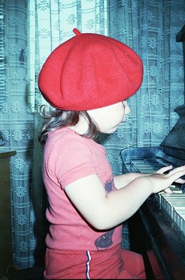 Vintage photo of a child wearing a red beret and pink sleeveless dress with blue whale patches, playing a vintage upright pia...