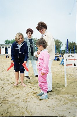 Four adults and a child pose on sandy terrain near a beachside playground. The adults wear 1970s-80s casual attire: jackets, ...