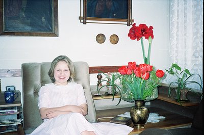 Vintage indoor portrait of a young girl in a white lace dress seated in a mid-century armchair, surrounded by framed art and ...