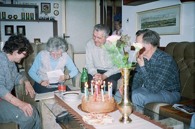 Family gathering in a mid-century living room celebrating a birthday. Centerpiece: multi-candle cake with floral decorations....
