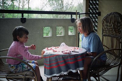 Indoor scene featuring two women playing a card game at a small round table draped with a striped blanket. The woman on the l...