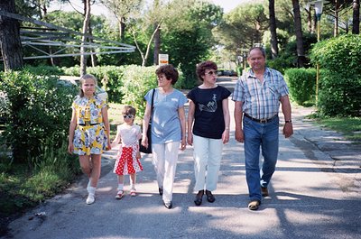 Family stroll in a lush, tree-lined park, 1970s-80s. Four adults and a child walk hand-in-hand on a paved path, dressed in re...