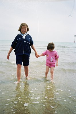Two children holding hands in shallow seawater, likely mid-1990s beach setting. Teen in navy zip-up tracksuit, toddler in pin...