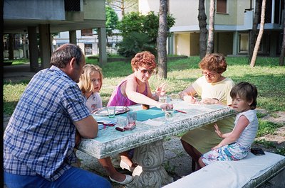 Family picnic scene at a concrete outdoor table in a landscaped residential area, likely 1970s–1980s. Five individuals—two ad...