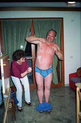 Vintage indoor scene featuring a man in blue undergarments and flip-flops balancing on a single foot, assisted by a woman in ...