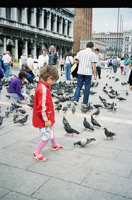 A child in a red Adidas tracksuit stands among a flock of pigeons in St. Mark’s Square, Venice. The 1990s-era clothing and br...