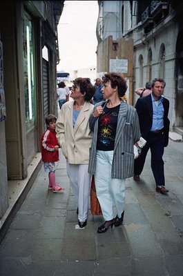 1970s street scene featuring three adults and a child in a European urban setting. Women wear bold patterned blazers and wide...