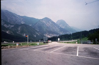 Empty alpine parking lot with forested mountains in background, likely a scenic viewpoint or trailhead. Concrete surface, min...