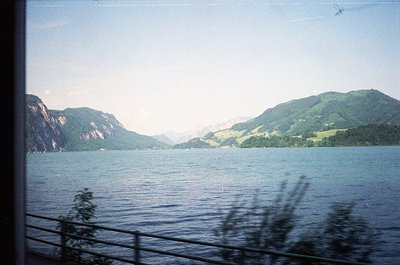Vintage train window view of a serene alpine lake framed by forested hills, captured through motion blur. Distinctive layered...