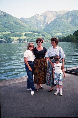 Three women and a child pose on a lakeside pier, framed by alpine scenery. The adult on the left wears a white T-shirt and su...
