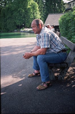 Man in casual 1970s attire sits on a park bench beside a serene lake, wearing a plaid shirt, jeans, and sandals. Lush greener...