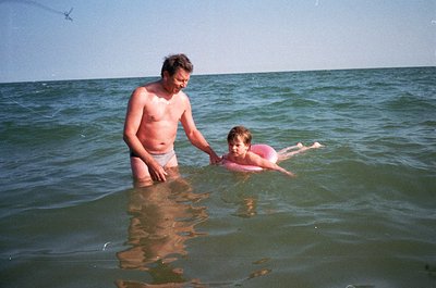 A man assists a young child in shallow seaside waters, mid-20th century. The adult wears loose swim trunks, while the child d...