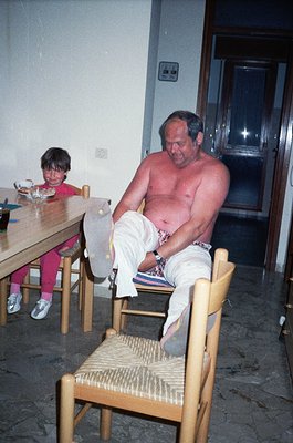 Mid-20th-century indoor scene: shirtless man in white shorts sits on woven chair, lacing shoes at wooden table. Young girl in...