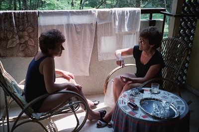 Two women seated in wicker patio chairs on a sunlit balcony, engaged in conversation. The woman on the left wears a sleeveles...