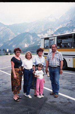 Family group photo in alpine setting, likely 1980s-1990s. Four adults and a young child pose near a yellow-and-blue bus in a ...
