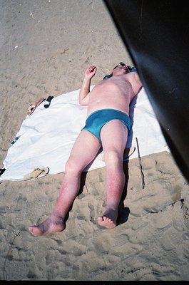 A person sunbathes on a sandy beach under a beach umbrella, wearing teal swim trunks. A white towel lies beside them. The ima...