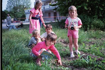 Five children and two adults in matching pink outfits explore a grassy, overgrown area near a residential building, likely a ...