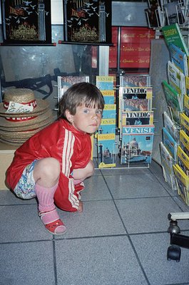 Child in 1980s-style Adidas tracksuit kneeling on tiled floor, surrounded by postcards featuring Venice landmarks. Stacked po...
