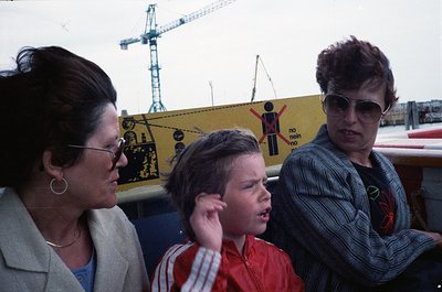 Three individuals stand near industrial construction site with safety signage. Woman on left wears glasses, beige blazer, and...