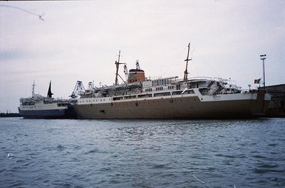 Mid-20th century passenger ship docked at port, likely –. White hull with red-orange funnel, multiple decks, and lifeboats vi...