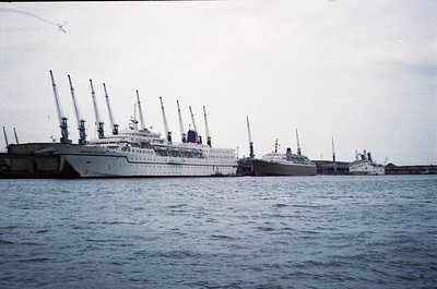 Mid-20th century ocean liner docked at port, likely 1950s–1960s. Multi-deck vessel with tall masts and white hull, flanked by...