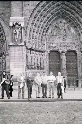 Gothic cathedral façade featuring intricate stone carvings, statues, and pointed arches. Group of men in 1960s-era clothing p...