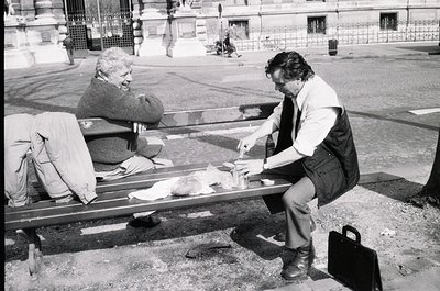 Two men share a meal on a park bench in an urban setting, likely mid-20th century. The seated man eats while the standing man...