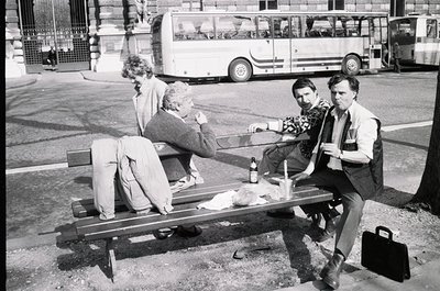Three men relax on a wooden bench in an urban setting, 1970s. One holds a bottle, another a briefcase; vintage bus and classi...