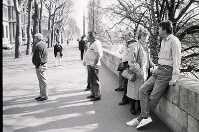Group of five adults in 1970s streetwear—men in button-downs, women in layered coats—standing on a wide urban promenade lined...