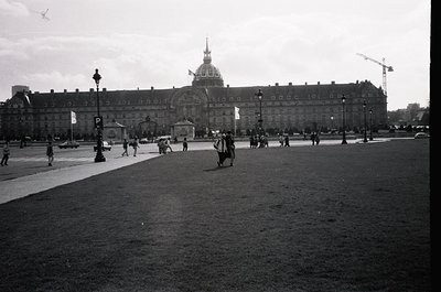 Neoclassical palace with domed roof and symmetrical façade, likely the Hôtel des Invalides in Paris. Crowded plaza with pedes...