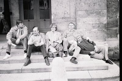 Four individuals pose casually on stone steps outside a stone-framed building, likely mid-20th century (1960s–1970s). Clothin...