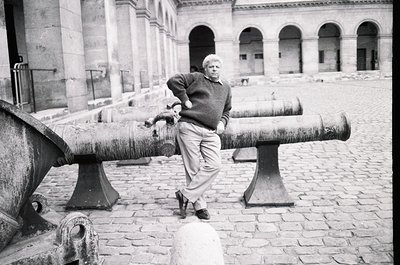 Man poses confidently atop 19th-century cannon in cobblestone courtyard, leaning on barrel with one hand. Architectural backd...