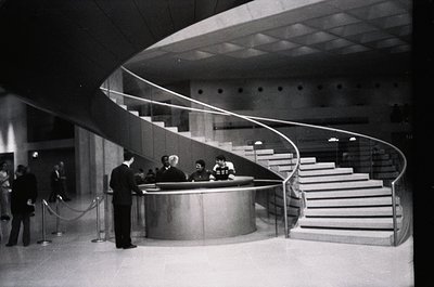 Mid-century modern reception desk with curved metal counter and sweeping staircase in a grand public building. Formal attire ...