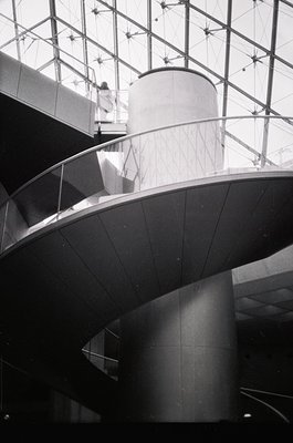 Modernist architectural interior featuring sweeping curved concrete ramps and cylindrical columns. Glass ceiling with structu...