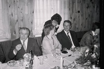 Mid-20th century formal gathering in a rustic wooden-paneled dining room. Six adults seated around a long table adorned with ...