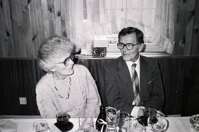 Mid-20th century indoor portrait: two adults seated at a table with glasses, plates, and a bottle. Woman in a patterned sweat...